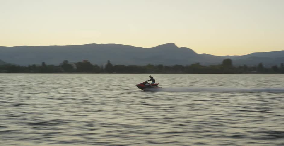 A man rides a jet ski through a large lake that runs parallel to a city and a mountain range