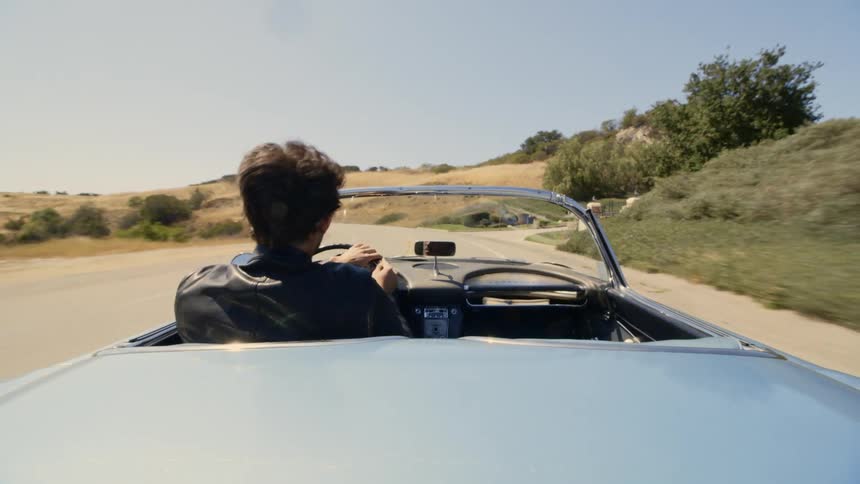 A man in a leather jacket driving a convertible along a road through hilly terrain on a sunny day