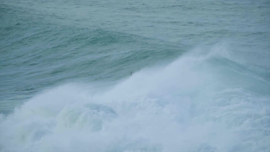 Male stands on surfboard and rides the wave as another male sits floating on his surfboard farther out into the ocean water