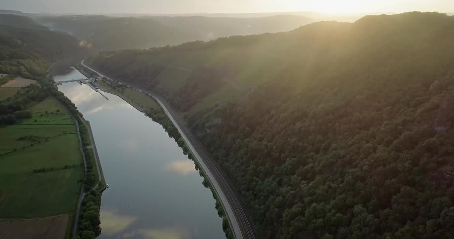 A river runs in a valley between farmland and a mountain covered in a forest as the sun sits on the horizon