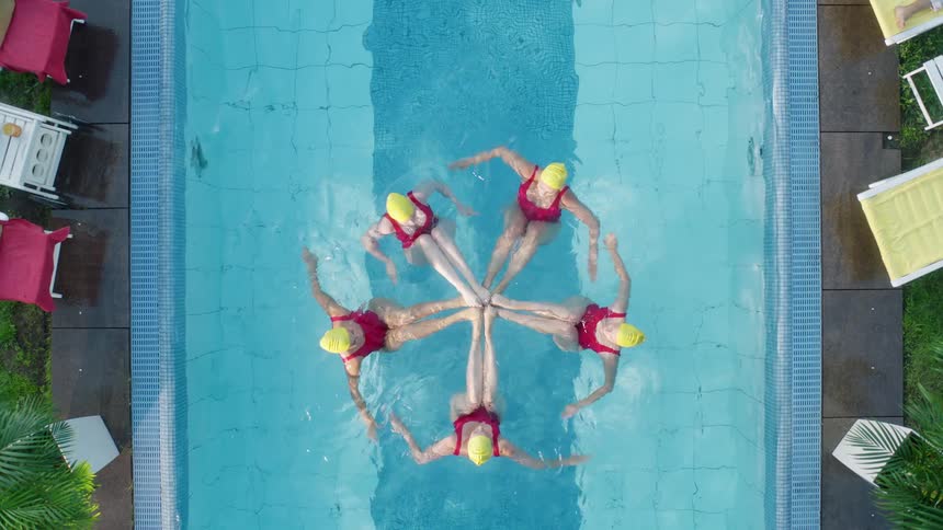 Five females wearing yellow swim caps and pink bathing suits perform a synchronized swim routine in a lap pool