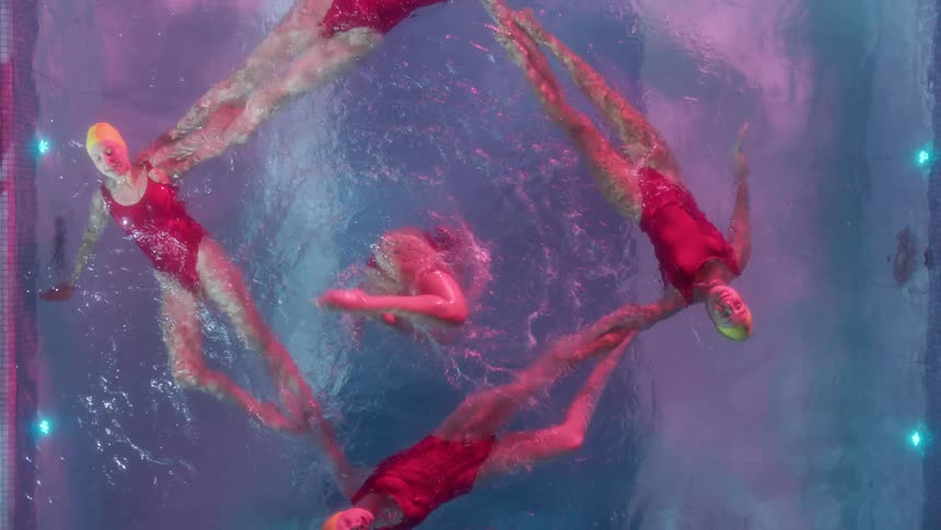 Four synchronized swimmers float and hold a square formation while another swimmer twirls and kicks with legs out of the water