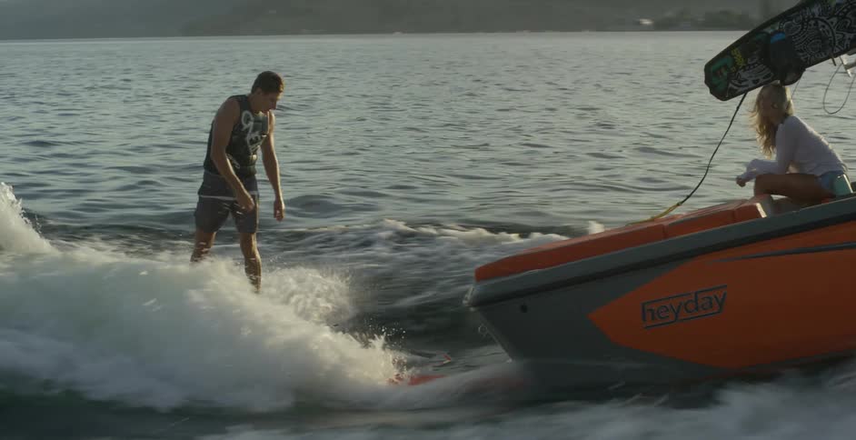 A woman sits facing the back of the boat watching a man wakeboard on a sunny day