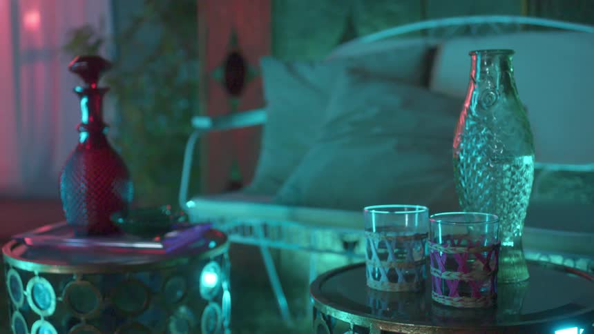 Glasses and decorative serving bottles on tables near a cushioned bench on an outdoor patio with soft colorful lighting