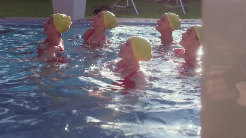 Five female synchronized swimmers wearing yellow swim caps and pink bathing suits come up out of the water at the same time and begin to move their arms while performing a routine in a swimming pool as the sun reflects off of the water