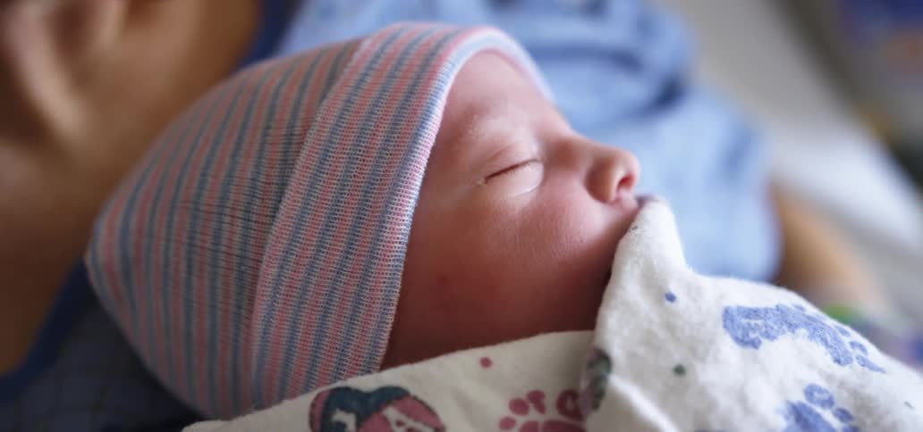 Newborn infant swaddled in a blanket sleeps on mothers chest as they lay on a hospital bed