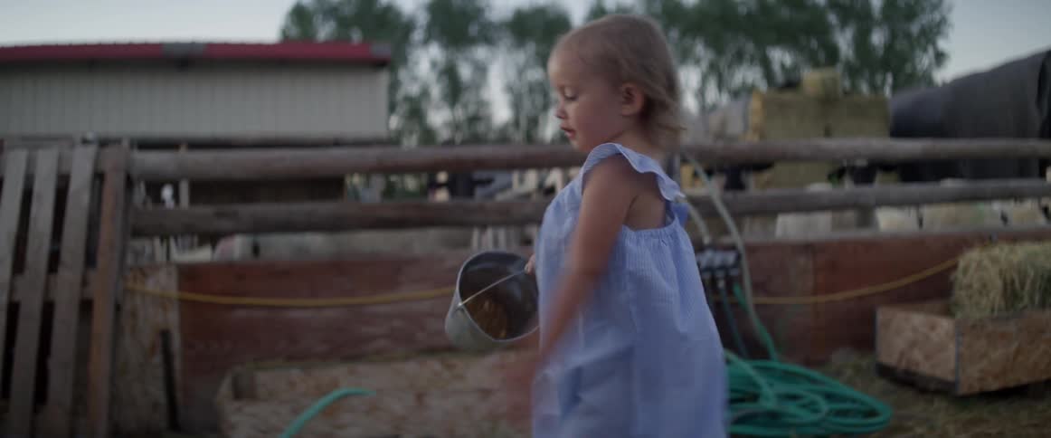Little blonde girl carrying bucket walking after mother to help on farm at sunrise