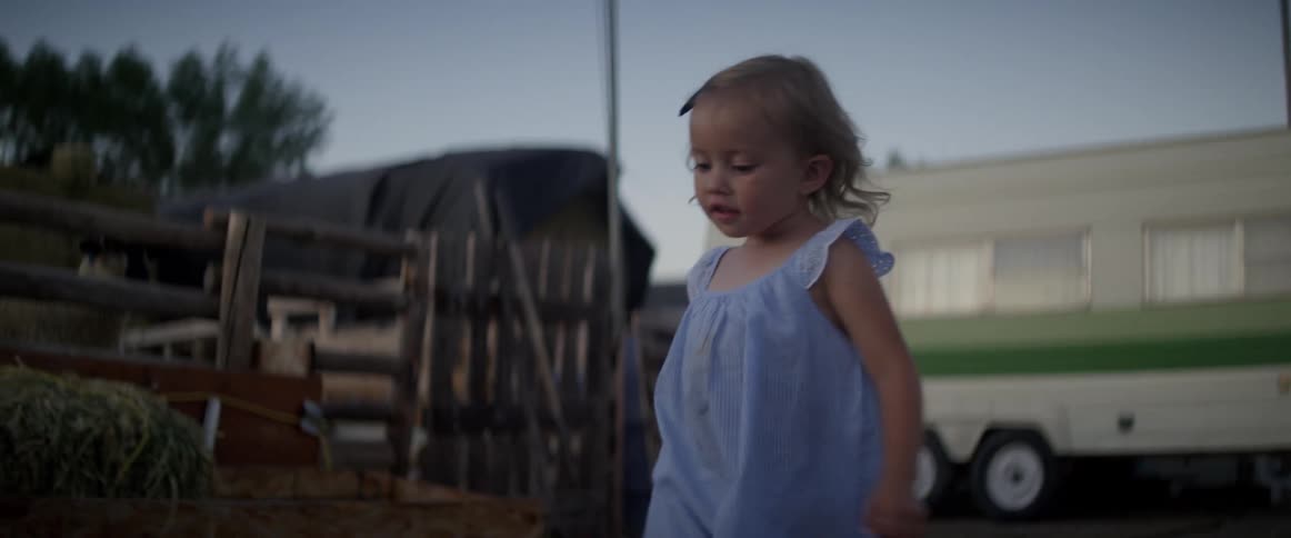 A little girl in a blue dress walks with a galvanized bucket near a green and white camper