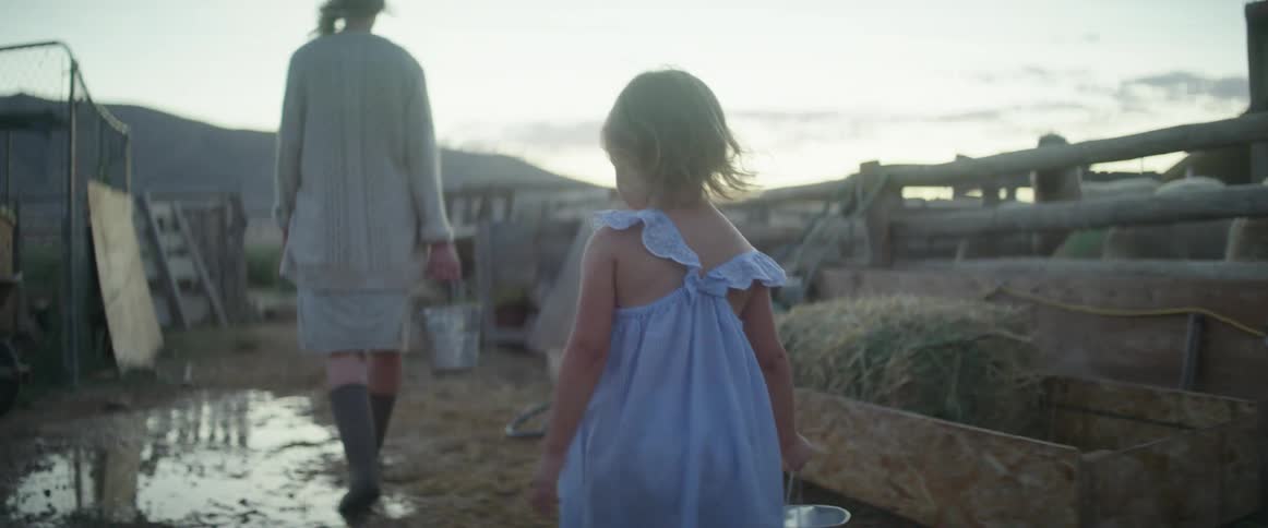 A young girl in rain boots jumps into a mud puddle while carrying a pail on a farm