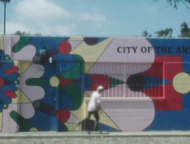 A white teen skateboards on a sidewalk next to a oddly painted building