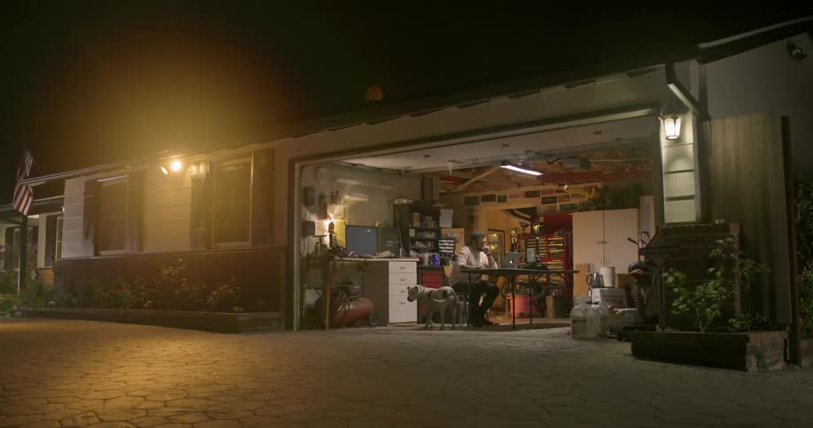 A man sitting in his garage working on his laptop reaches down and pets his dog