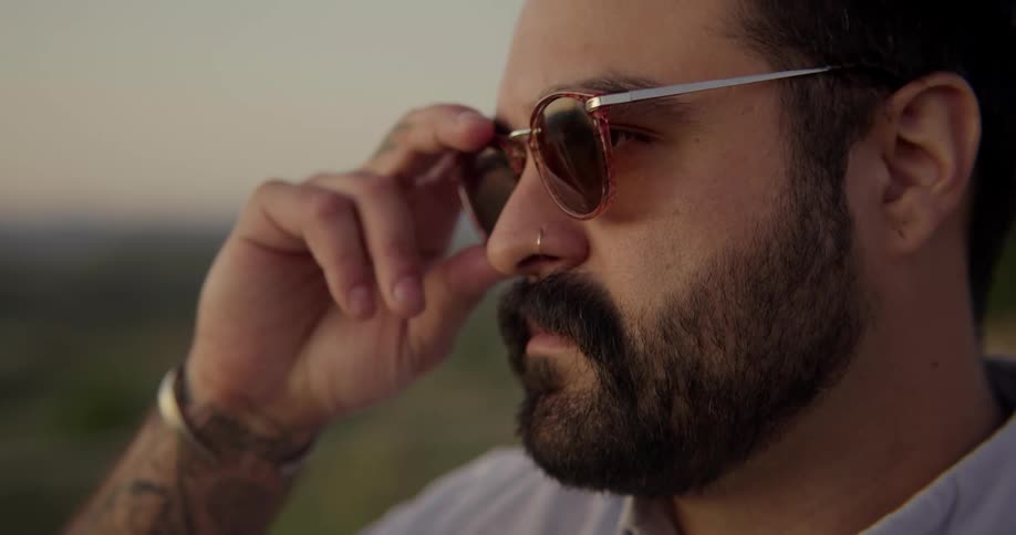 A bearded man puts on sunglasses while standing outdoors