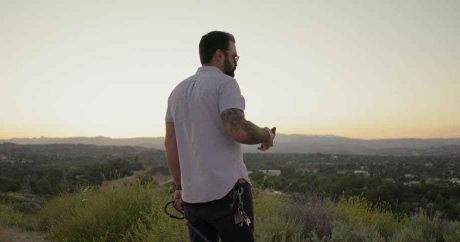 Tattooed man with sunglasses and beard stands and talks on the smartphone on a hill at dusk