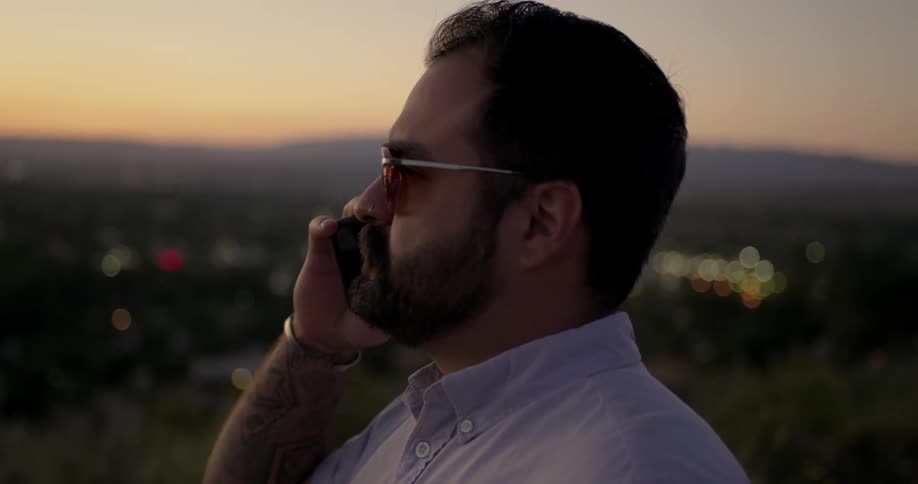 A tattooed middle aged man rising a smartphone to his ear and speaking into it as he looks out over the lights of the city below