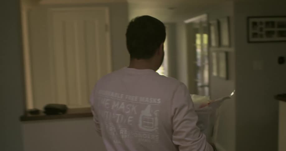 A white man picks up a plastic bin filled with products and carries it outside of his home