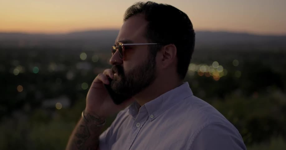 Man with beard and sunglasses talks on a smart phone while walking on a hill at dusk