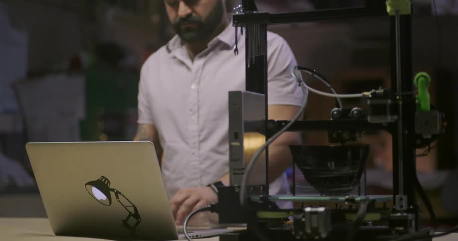 A man walks to a desk with a 3d printer on it and types on a laptop