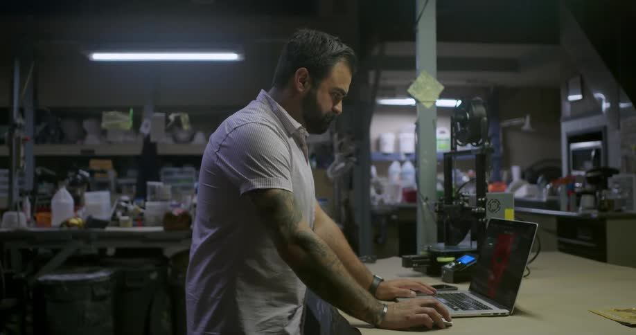 A man walks over to the laptop by a 3d printer