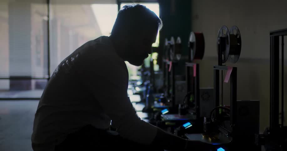 A man is sliding on a chair checking the row of 3d printers