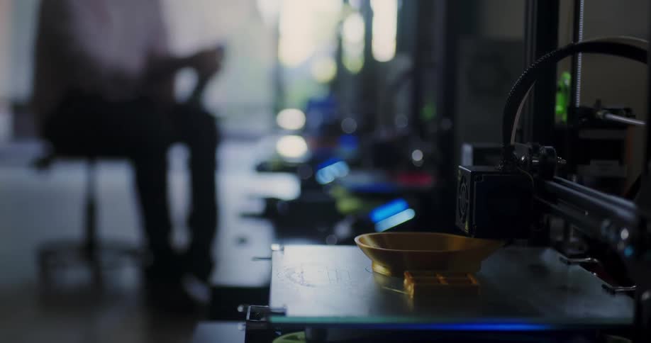 A 3d printer makes a golden bowl while a male tends to other machines in the distance