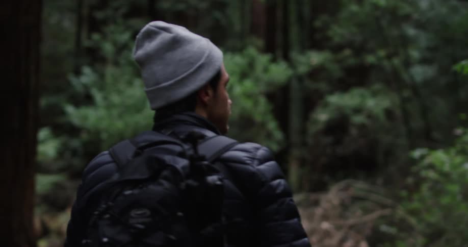 Male carrying a tripod in his backpack hikes through the woods and looks around while walking