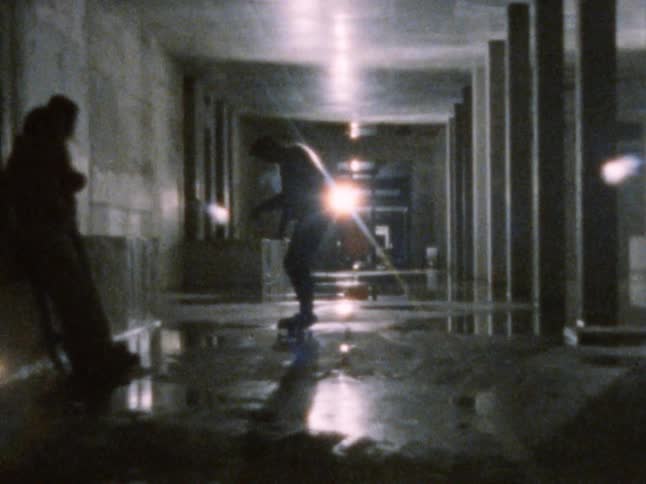 Two males skateboard inside a dank building while another male leans against a wall watching