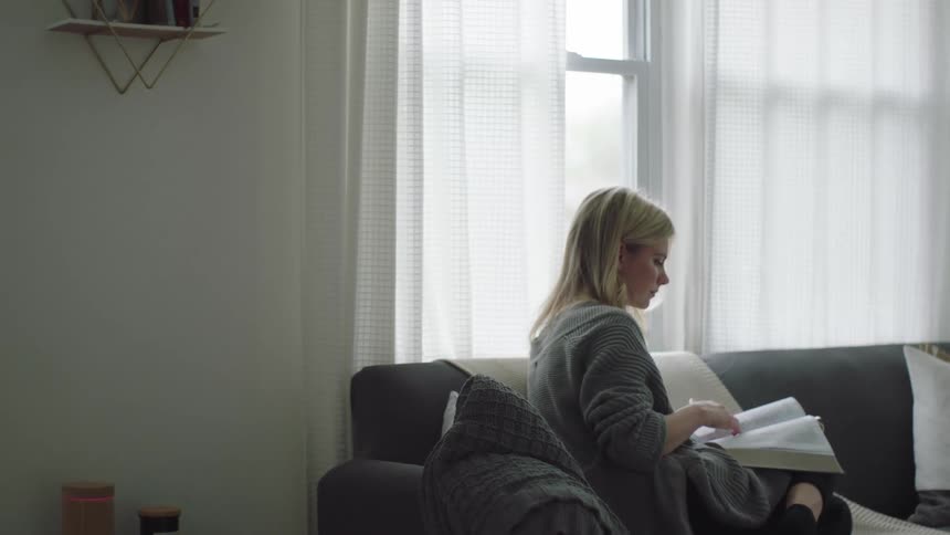 A blonde woman sitting on sofa leafs through a book in the living room at home