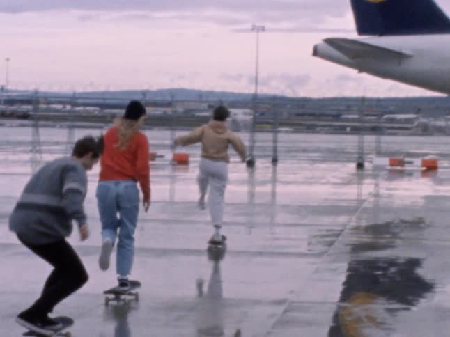 Group of skateboarders ride boards across wet tarmac near airplane