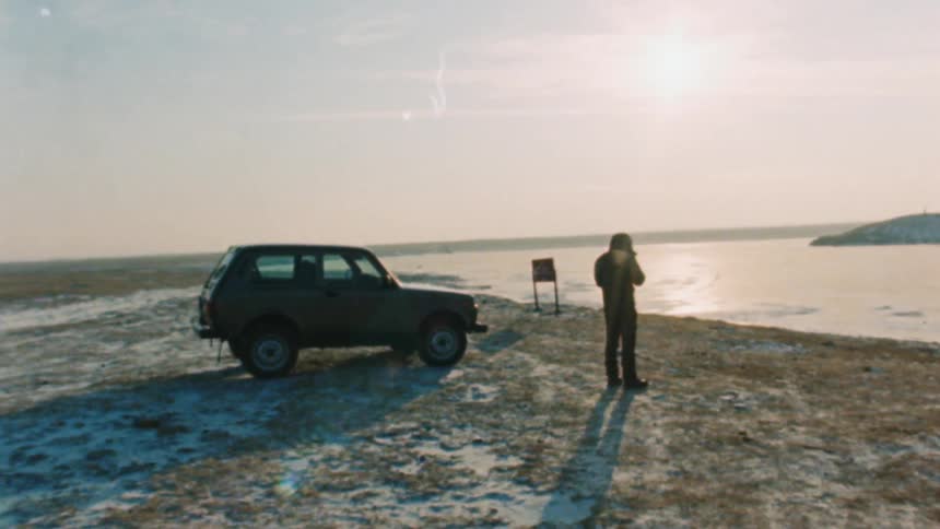 A man standing next to a car parked in the field next to a frozen lake
