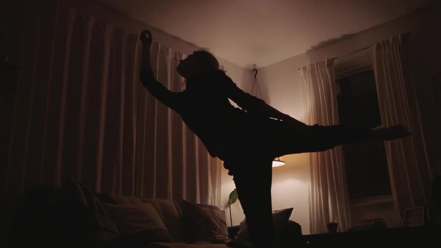 A woman stretches upward into a ballet dance pose in a dark living room