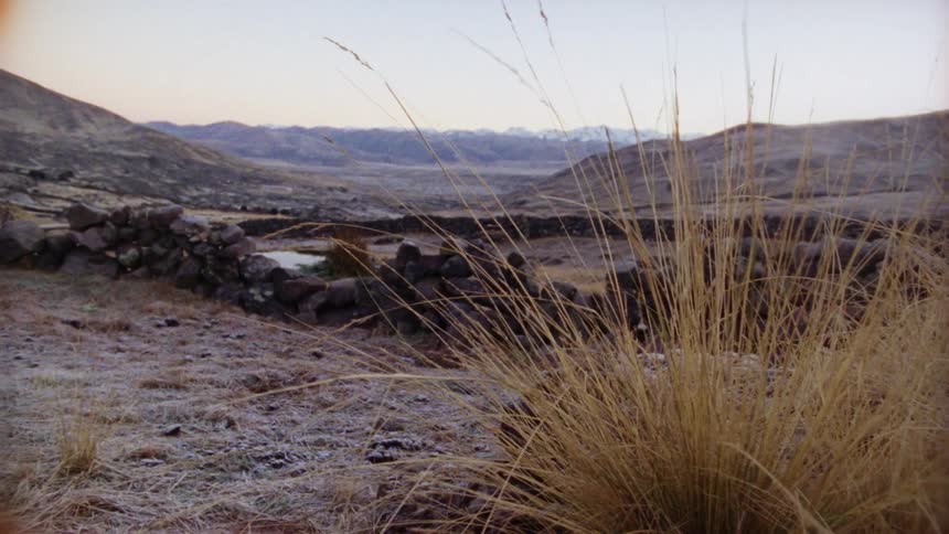 Clumps of grass dot ground near stacked stone half wall with mountains on horizon