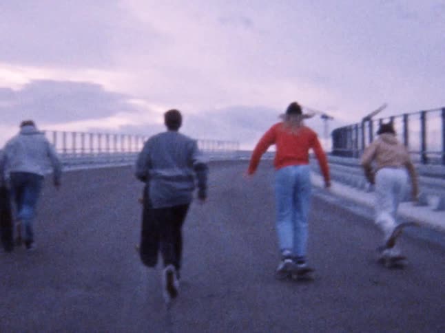 Four teens walking along the asphalt and then jump on their skateboards and start riding