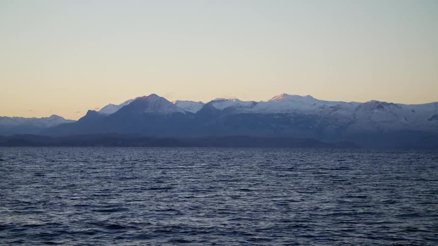 Ocean waves ripple with snow capped mountains on shore at sunset