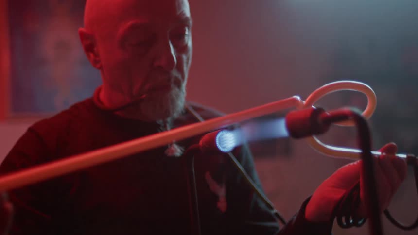 An elderly white man twists a tube through a blue flame
