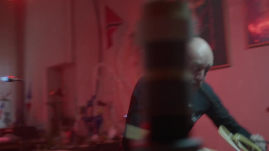 A bald old man heats and bends a tube of glass with a lampworking torch before moving it to a table to continue shaping it