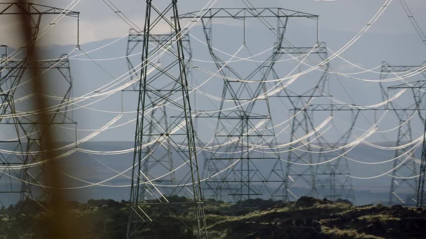 Rows of transmission towers and cables dominate a hilly nature area