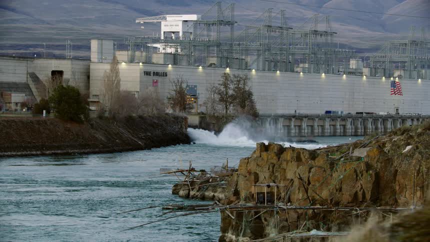 Water gushes into river flowing past power station hydro electric dam with mountains in background