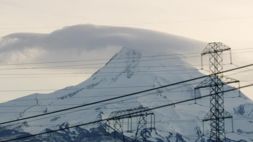 Transmission power lines in front of a large mountain volcano with deep snow deep in the winter at sunset with a train passing in the foreground at the end of the shot