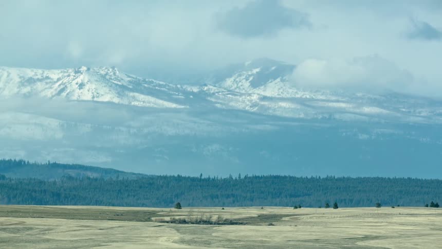 A vast winter swept rural landscape from the point of view looking out the window of a driving vehicle at huge snow covered mountains