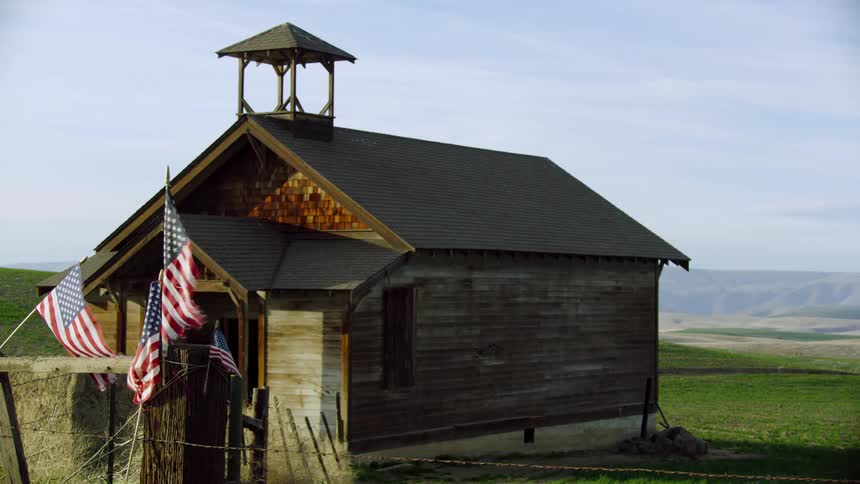 A historical and abandoned school house in rural America with several American flags waving in the afternoon breeze with a large green meadow in the background