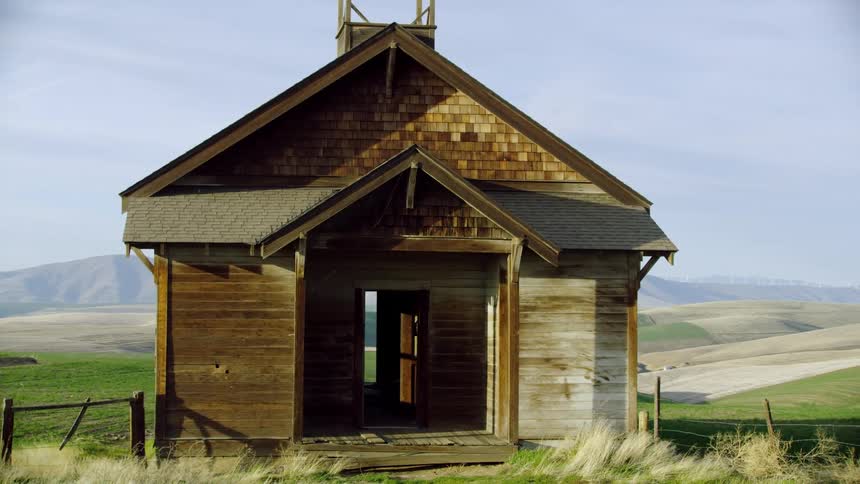 A rural and old abandoned wooden building atop a hill in a vast rolling countryside as the camera dynamically moves past it from the POV of a passenger looking out the window of a farm truck