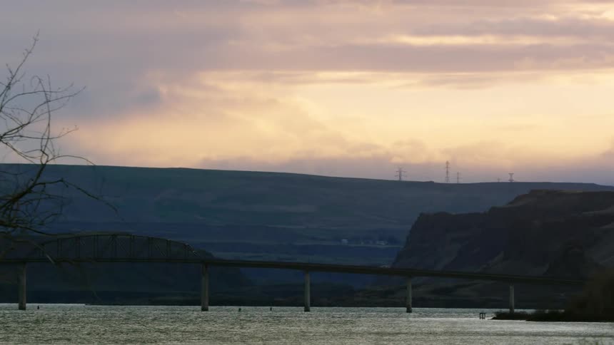 Shot looking back at a large river bridge spanning the Columbia River at sunset with leafless t in the foreground