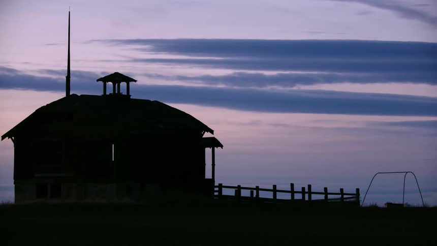 Tight shot of an old abandoned school house silhouetted agains pink and purple blue hour skies as the camera rotates around the old building