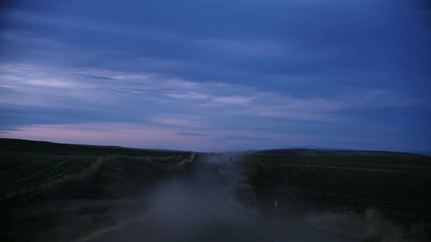 Ideal blue hour and pink sky shot looking out the back window of a pickup truck as it drives home on a dirt road with dust flying high in the sky after a long day of work