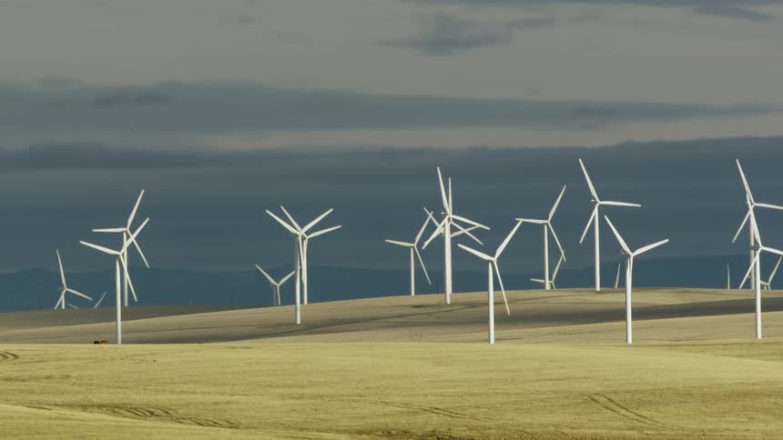 Low flying aerial shot of white windmills pleasantly rotating in front of dark afternoon clouds generating clean renewable green energy for electric utility customers