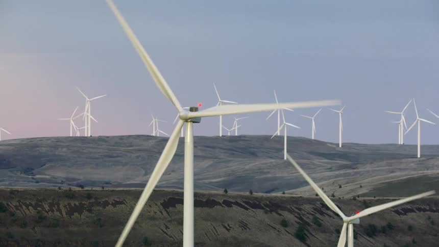 Medium shot titling up to frame a prominent wind turbine spinning in the foreground with many more white turbines turning in the background generating renewable electricity