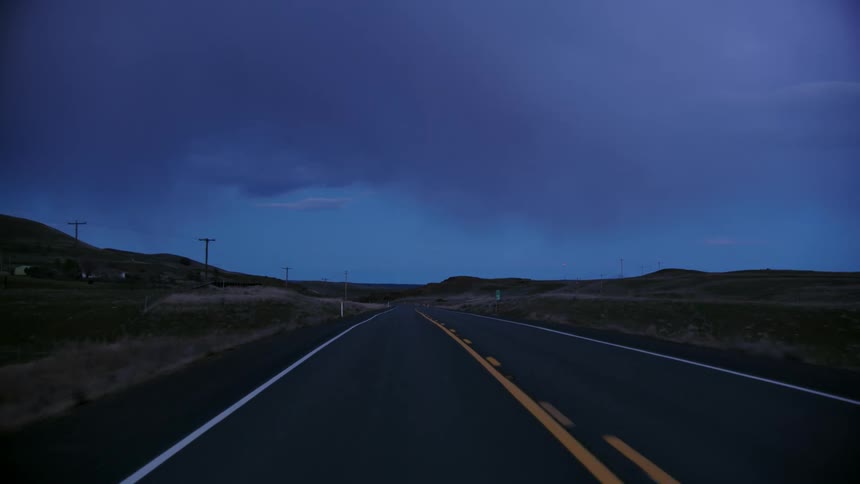 Looking back at a carless highway after the sun has set with power poles on one side of the road and open meadow on the other side
