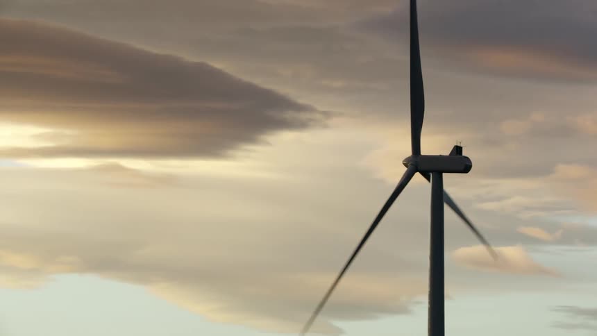A silhouetted wind turbines is followed by the camera in this low altitude aerial with beautiful sunset clouds in the background