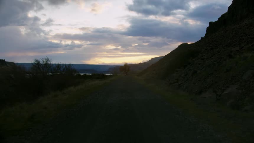 Looking back on a dusty rural dirt road at sunset as the vehicle drives along a craggy road with a large river and cliff