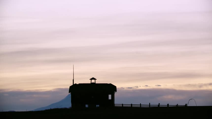 Wide shot of a silhouetted old school house against a vast sunset sky and Mount Hood in the background as the car passes by the rural landscape